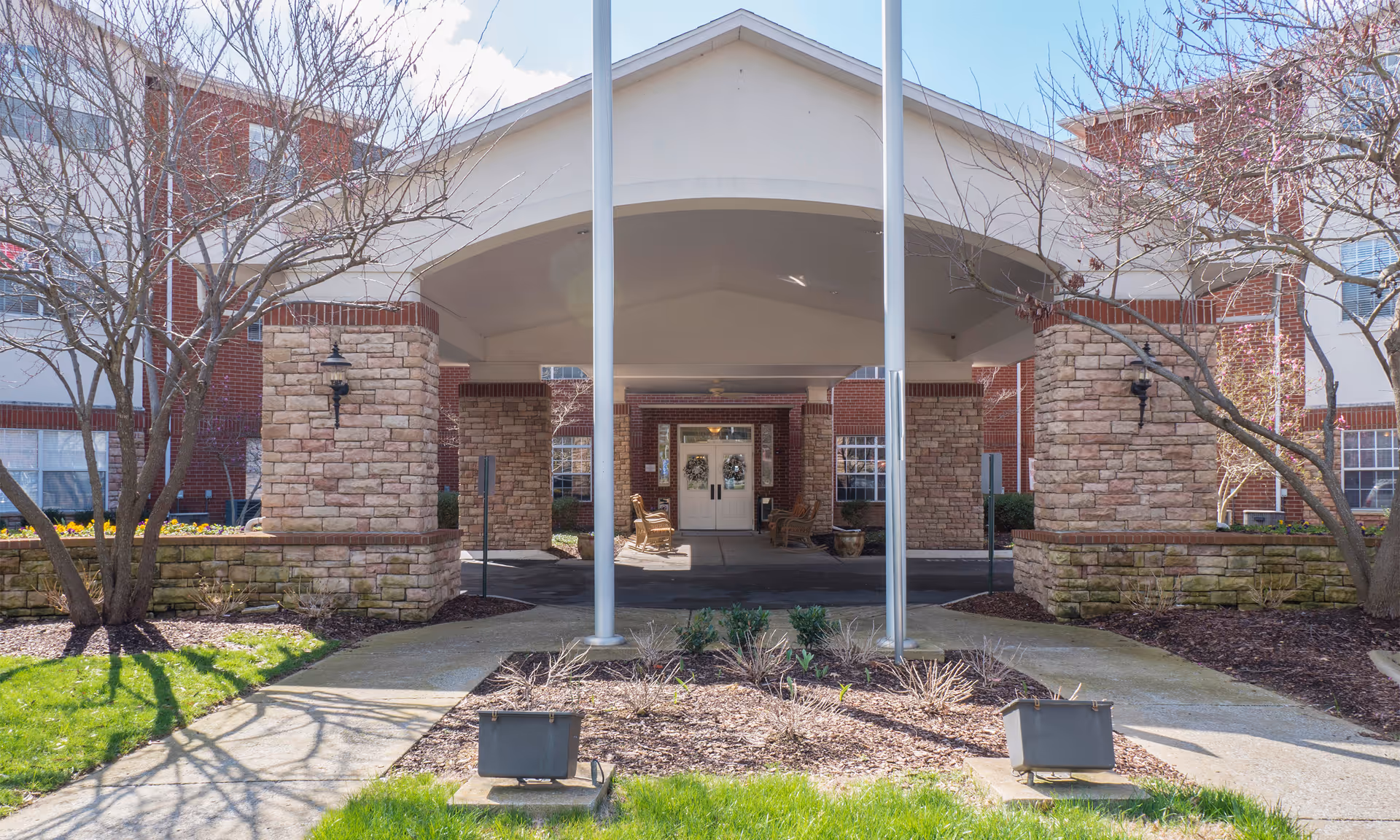 Front entrance of a senior living facility with a covered driveway supported by stone pillars, two flagpoles in front, leafless trees on either side, and a pathway leading to double doors decorated with wreaths.