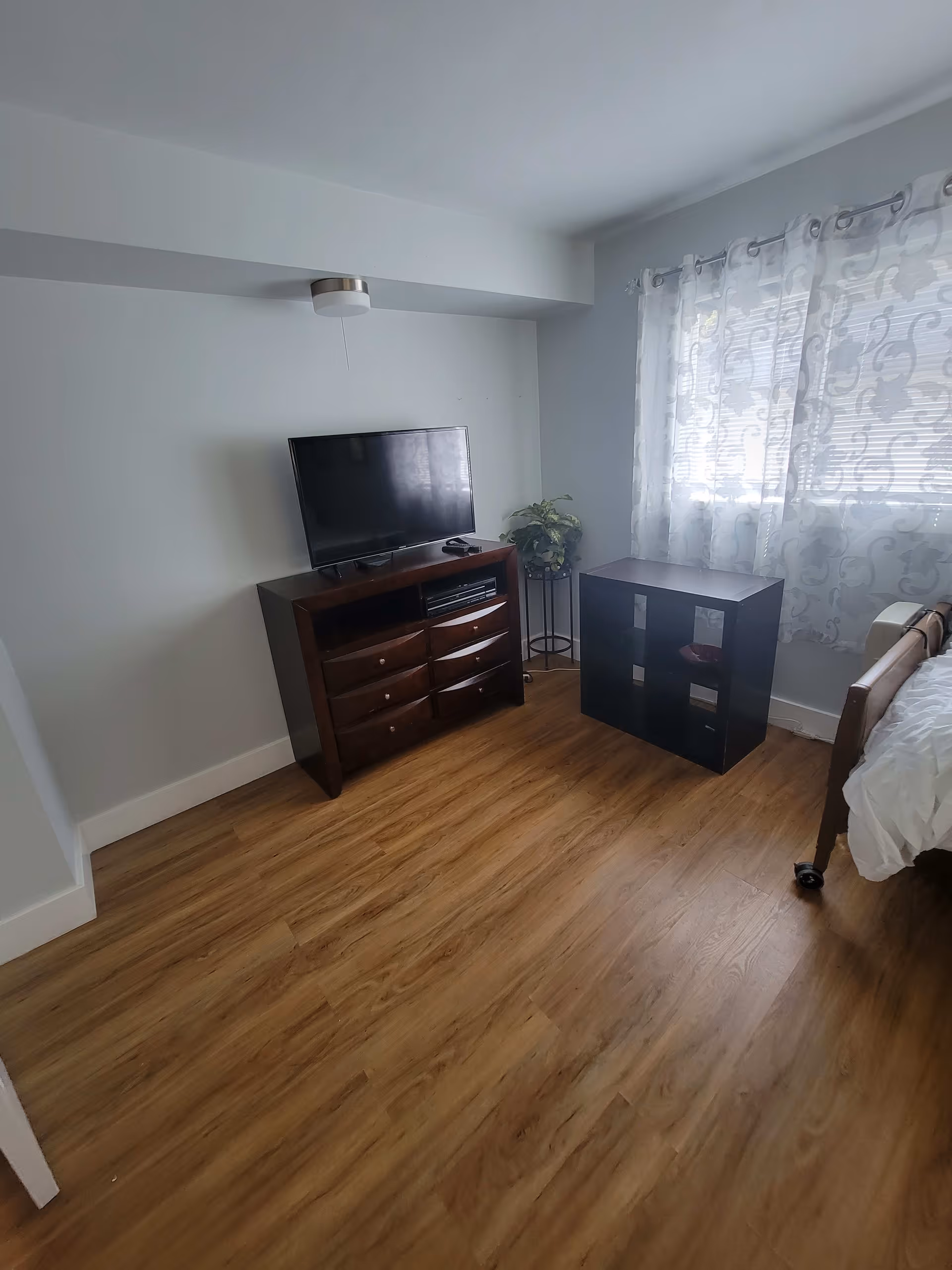 A small room with wooden flooring featuring a dark wooden dresser with a flat-screen TV on top, a small black shelving unit, a potted plant on a stand, and a bed partially visible on the right side near a window with sheer patterned curtains.