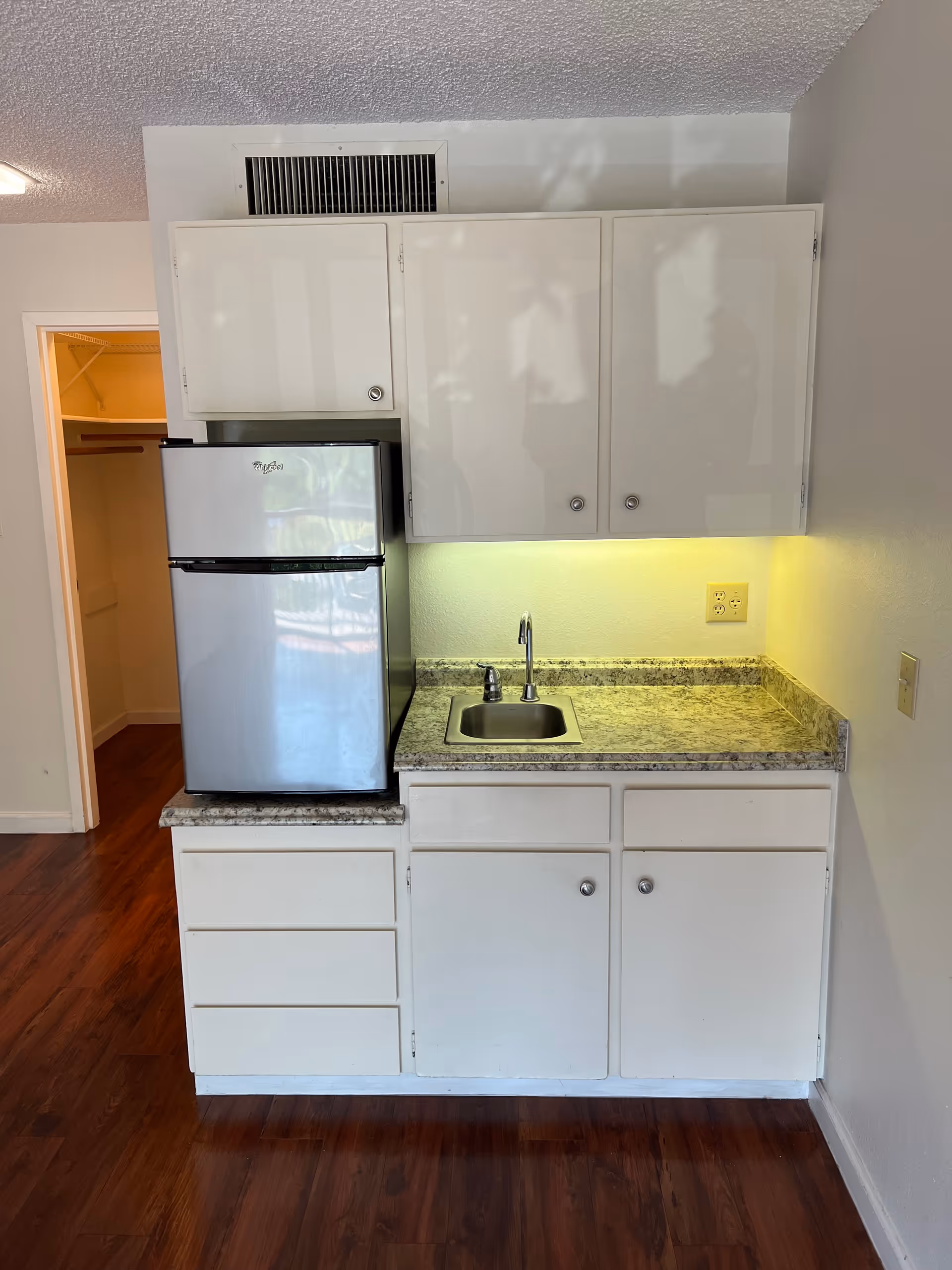 Small kitchenette with white cabinets, a compact refrigerator, a sink and a granite-look countertop against a light wall and wood floor.