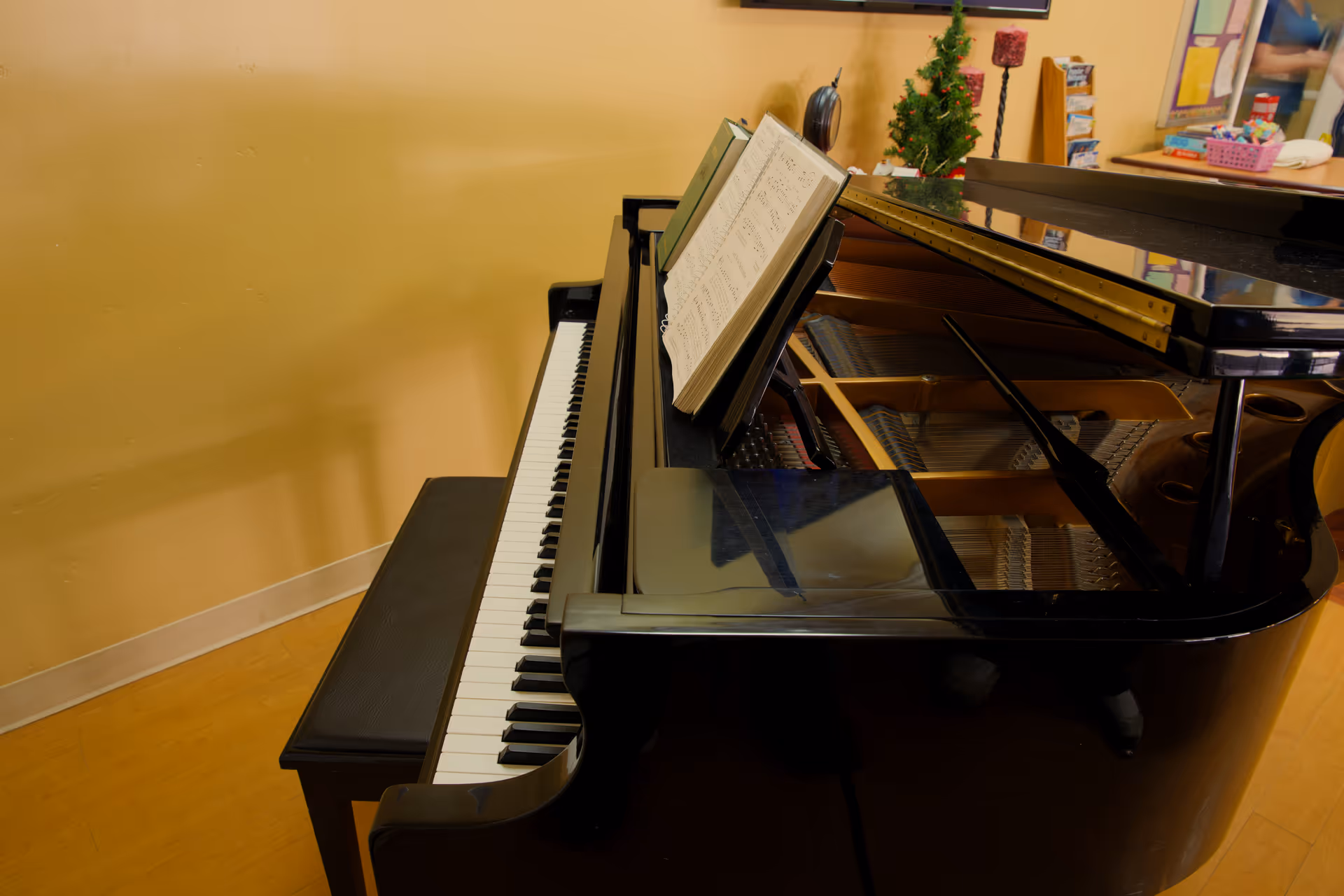 A black grand piano with an open lid and sheet music on the music stand, placed against a yellow wall with a piano bench in front. In the background, there is a small decorated Christmas tree and some brochures on a counter.