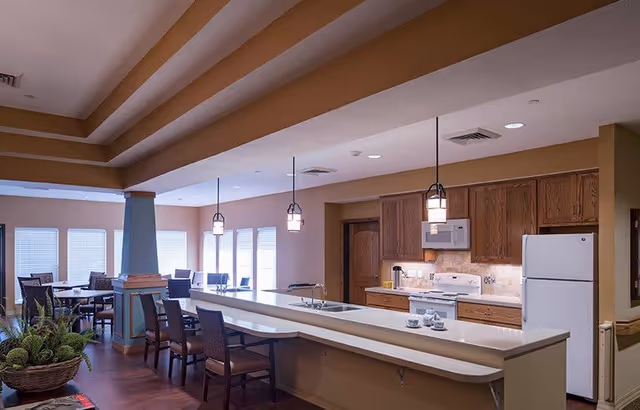 Open communal kitchen and dining area with a long island bar, pendant lights, wooden cabinets, and several dining tables and chairs by windows.