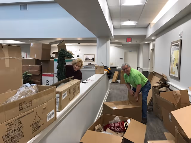 Two people unpacking and organizing boxes in a hallway of a facility. The hallway has white walls, a gray carpet, and overhead fluorescent lighting. Various cardboard boxes, some open and some closed, are scattered around the area.