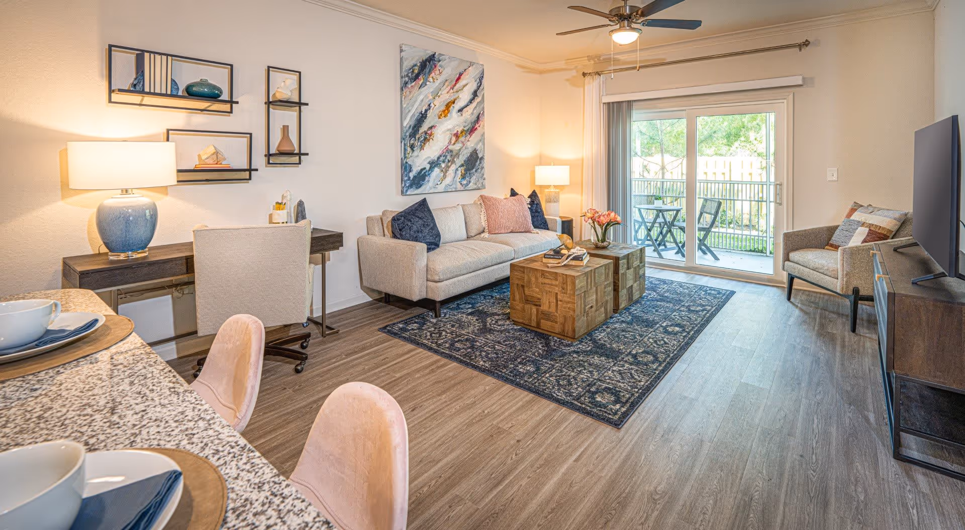 A bright and modern living room featuring a beige sofa with decorative pillows, a wooden coffee table on a patterned rug, an armchair, a flat-screen TV on a wooden stand, a desk with a lamp and chair, and sliding glass doors leading to a small outdoor patio with a table and two chairs.
