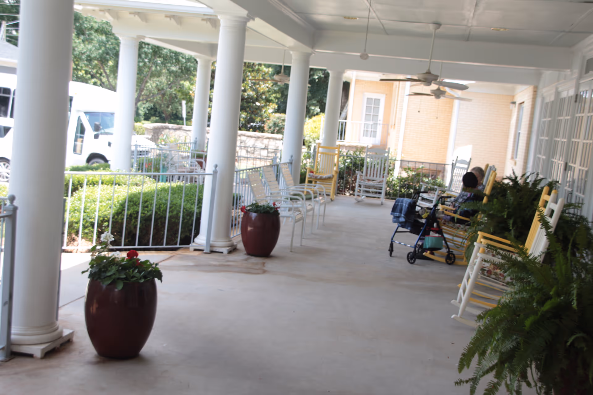 A covered outdoor porch area with white columns, several yellow and white rocking chairs, potted plants, and a person sitting on a rocking chair next to a walker. The porch overlooks greenery and a stone wall.
