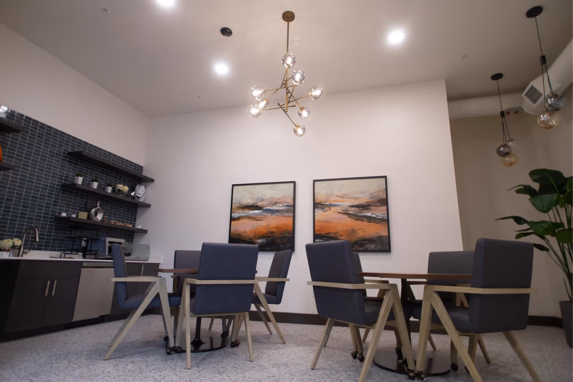 Modern communal dining area with tables, gray upholstered chairs, a small kitchenette and two landscape paintings on a white wall.