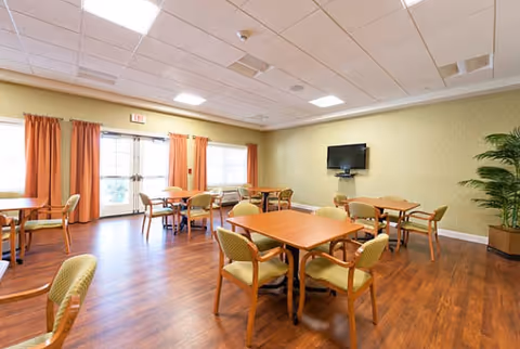 A bright dining room with wooden floors, several square wooden tables each surrounded by four cushioned chairs, large windows with orange curtains, a wall-mounted flat screen TV, and a potted plant in the corner.