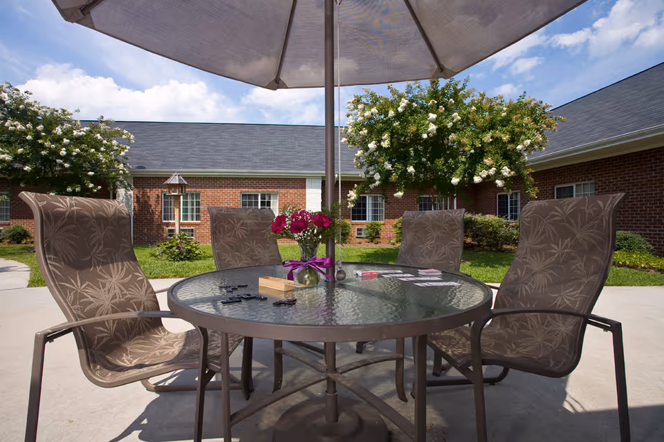 Outdoor patio area with a round glass table and four patterned chairs under a large umbrella. On the table, there is a vase with purple flowers, playing cards, and dominoes. In the background, there are brick buildings, green grass, and flowering trees under a partly cloudy sky.
