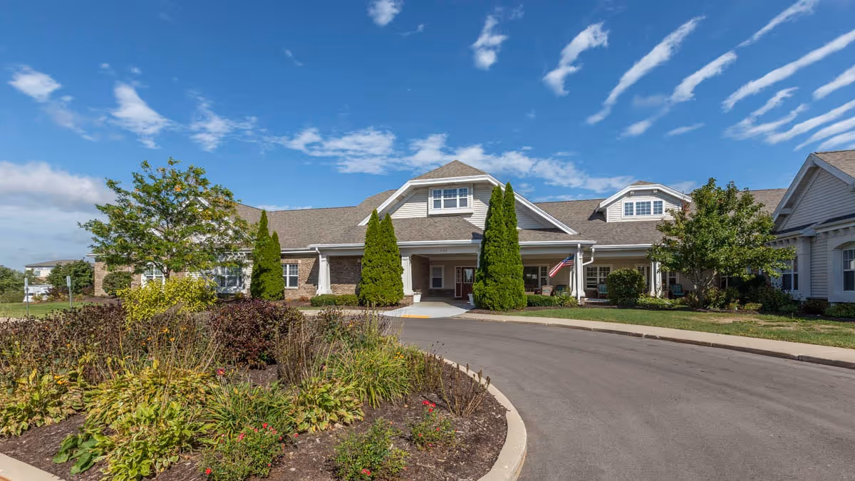 Front exterior view of a single-story senior living facility building with a covered entrance, surrounded by landscaped greenery and trees under a blue sky with scattered clouds.