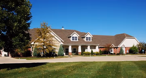 Exterior view of a single-story building with a sloped roof, dormer windows, and a covered entrance. The building is surrounded by trees and a well-maintained lawn under a clear blue sky.