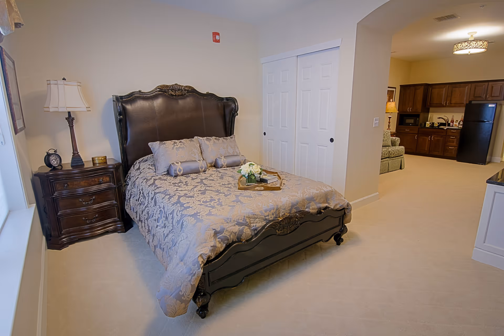 An elegant bedroom with a large ornate bed, nightstand and lamp, and a view into a kitchenette and sitting area.