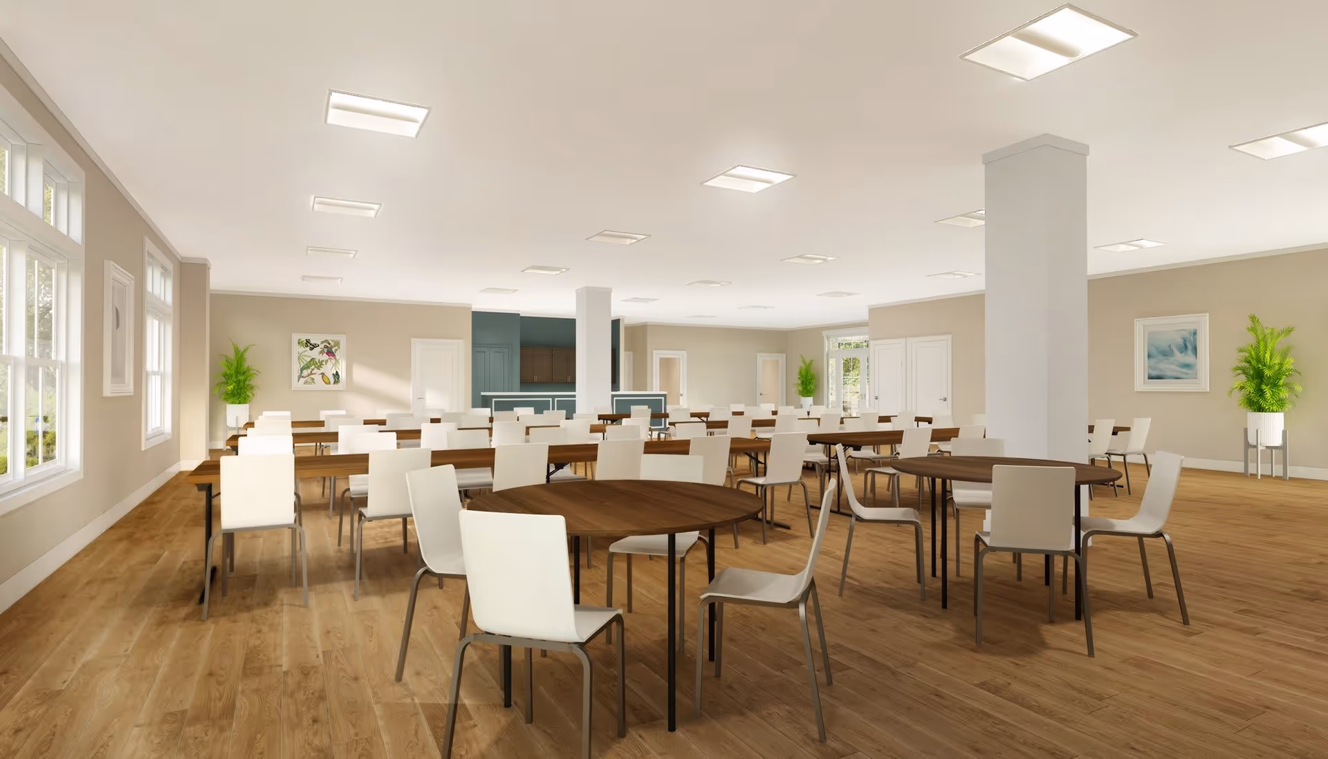 A spacious, well-lit dining room with multiple round and rectangular wooden tables surrounded by white chairs. The room has light-colored walls, large windows letting in natural light, wooden flooring, and some green plants in white pots placed near the walls. There are framed artworks on the walls and ceiling lights evenly distributed across the ceiling.