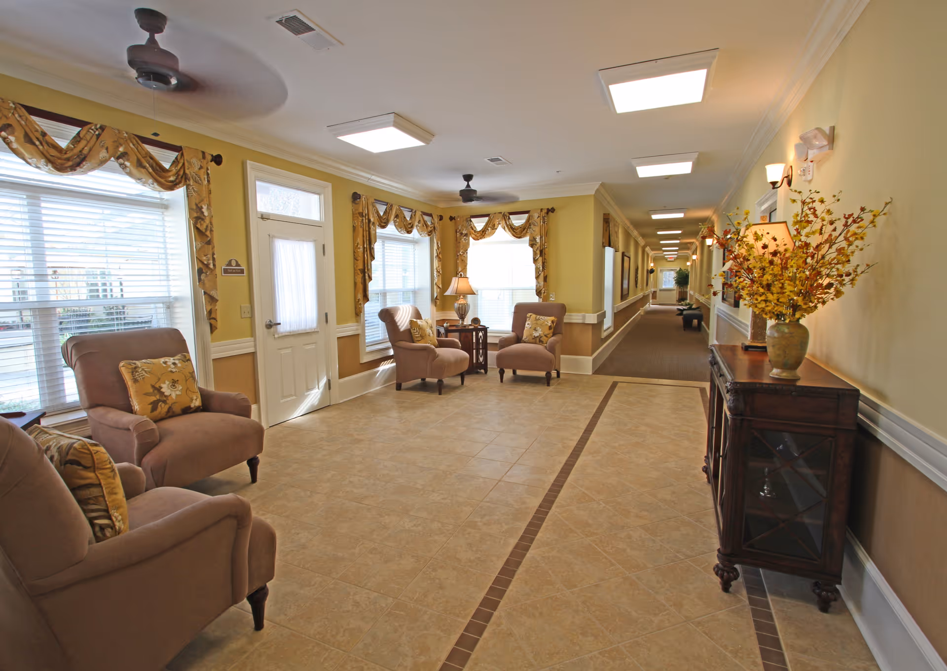 A bright and spacious senior living facility common area with beige tiled flooring and light yellow walls. The room features several comfortable armchairs with floral cushions, a wooden side table with a lamp, large windows with decorative valances, ceiling fans, and a wooden cabinet with a vase of yellow flowers. A long hallway extends from the room, lined with more seating and artwork on the walls.