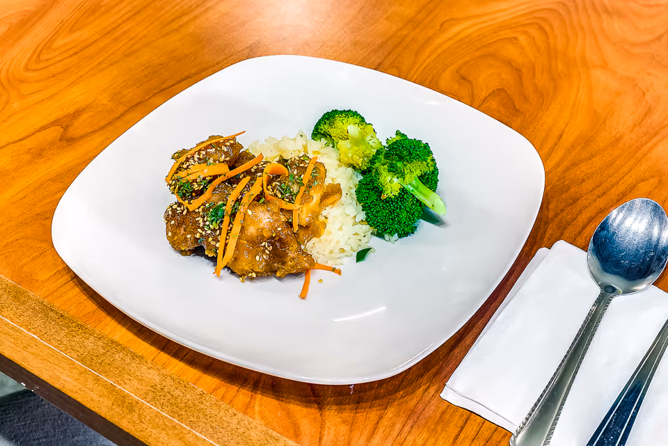 White plate with rice, broccoli and sesame-topped meat garnished with shredded carrots on a wooden table next to a spoon and napkin.