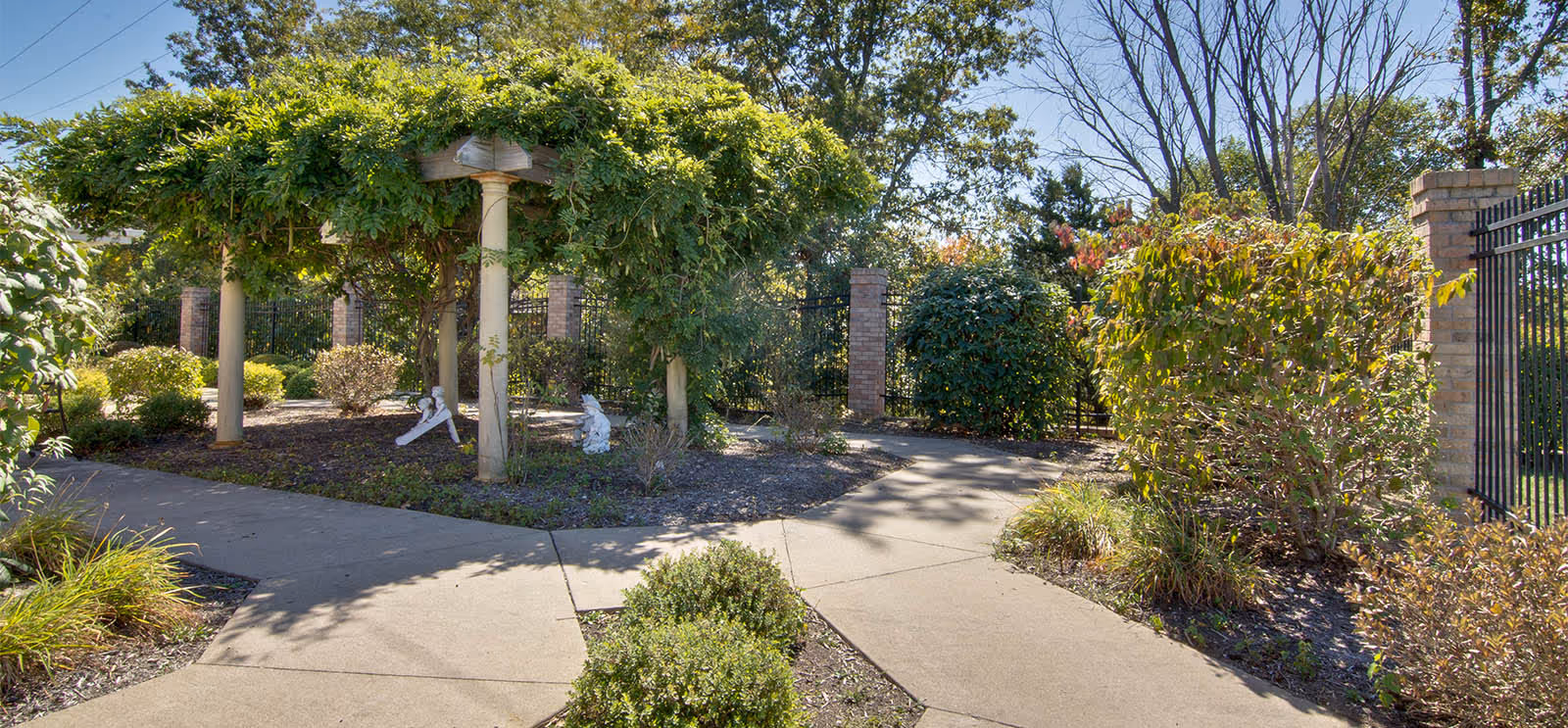Sunlit landscaped courtyard with a pergola, paved walkways, shrubs, and decorative statues near a brick-and-iron fence.