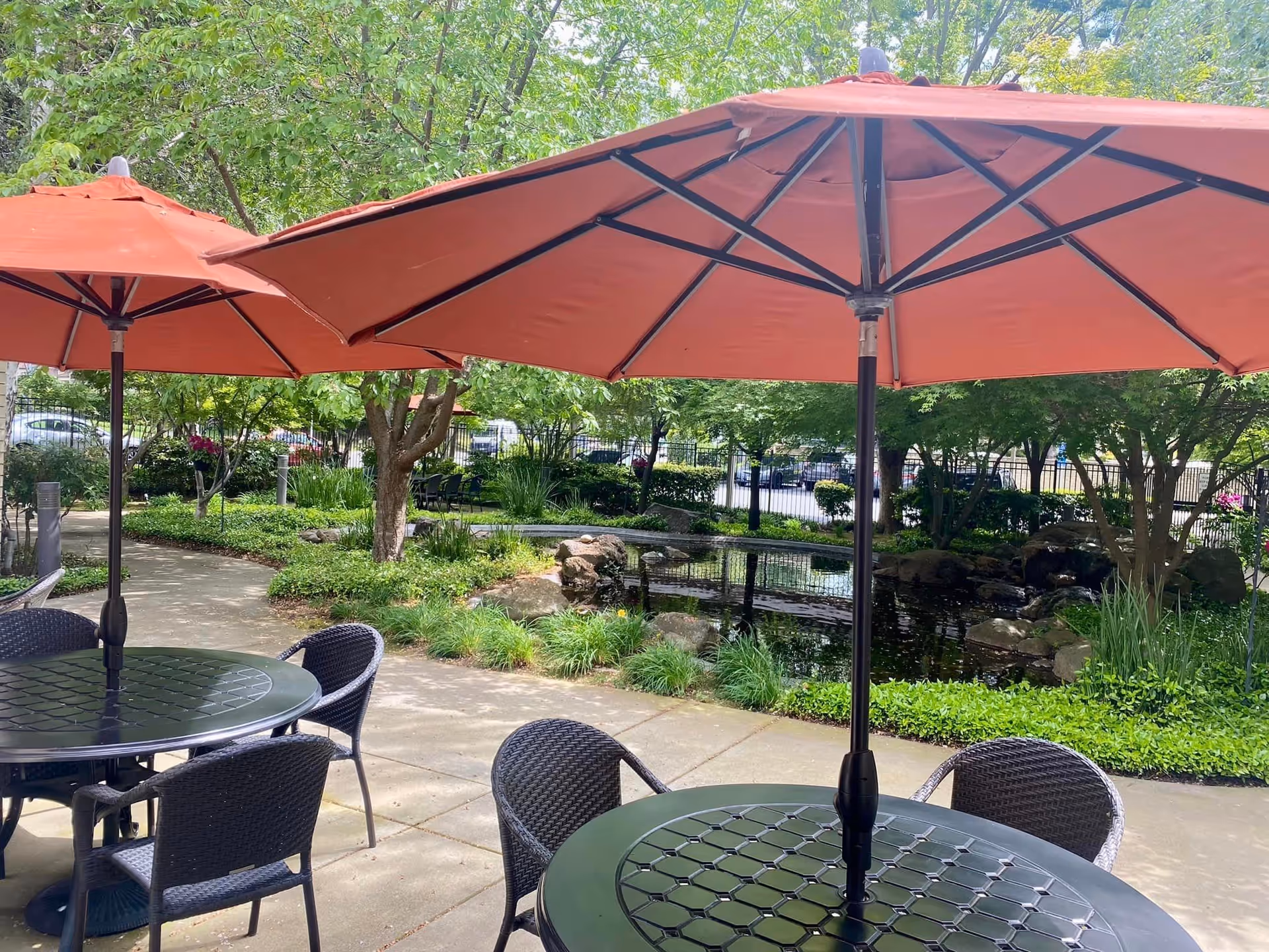 Outdoor patio area with round black tables and wicker chairs under large orange umbrellas. The patio overlooks a landscaped garden with a small pond, rocks, and lush green trees and shrubs.