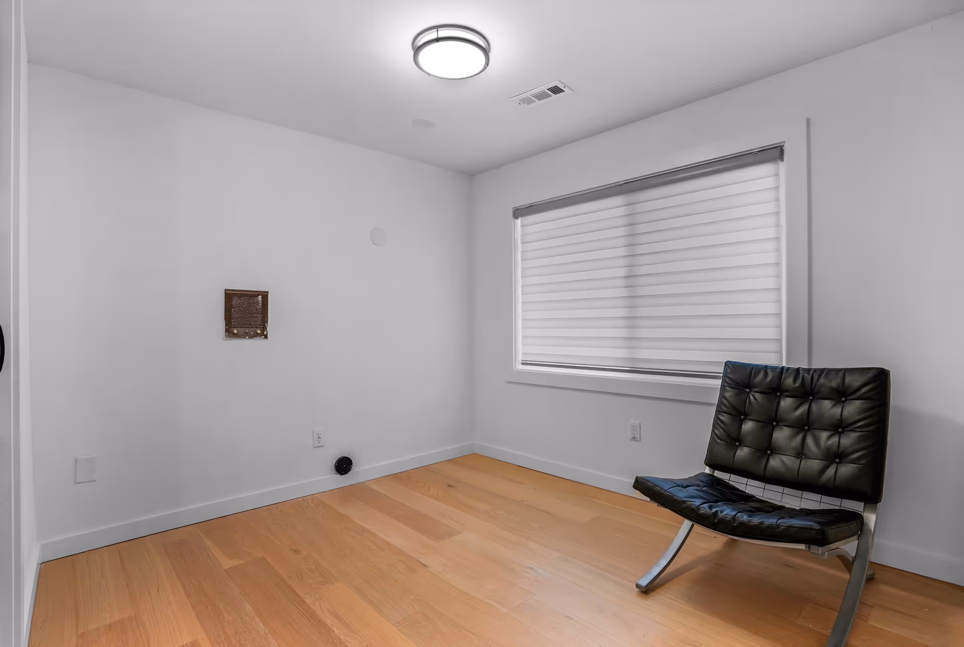 A small, empty room with light wooden flooring, white walls, and a single black modern chair with a metal frame. There is a window with a white horizontal blind on the right wall and a round ceiling light fixture. The room appears minimalistic and clean.