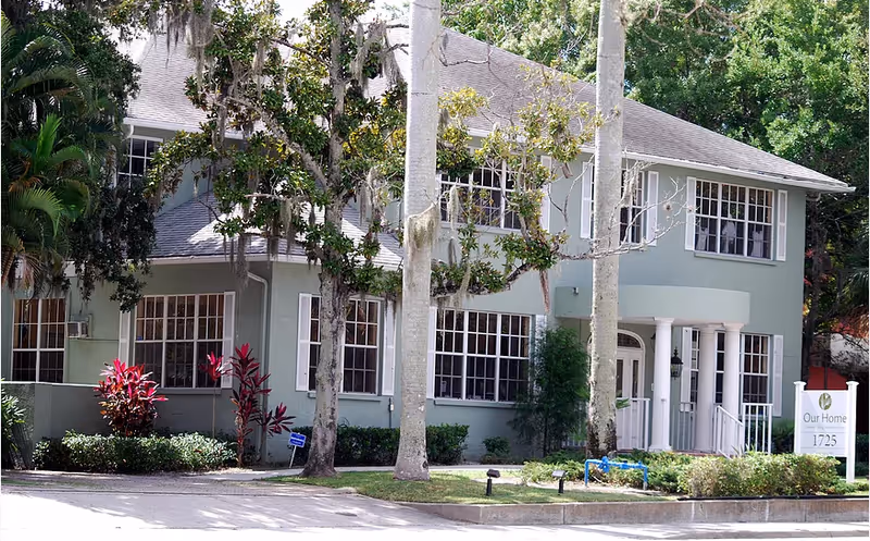 Two-story green senior living building with large windows, trees, and a front entrance sign.
