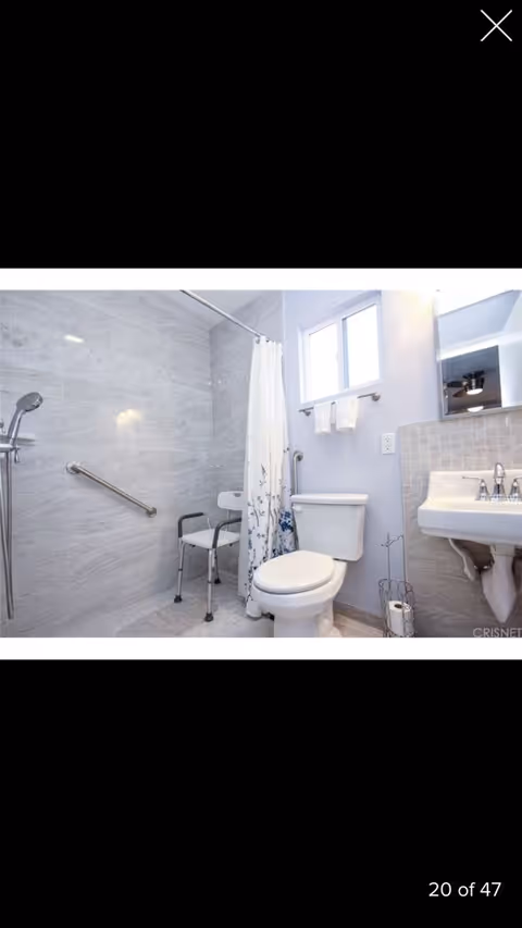 A clean, accessible bathroom with a walk-in shower featuring a handheld showerhead and a shower chair. There is a grab bar on the shower wall, a white toilet with a towel rack and two towels above it, a white sink with a mirror above, and a toilet paper holder on the floor. The walls are light-colored with tile accents.