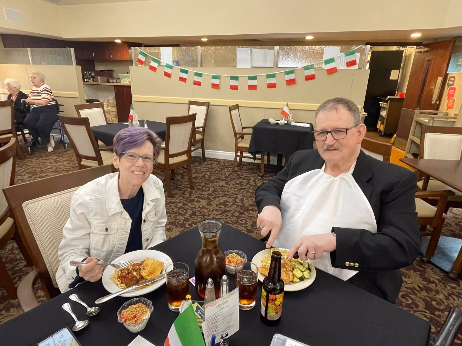 Two elderly people sitting at a dining table in a senior living facility, enjoying a meal. The man on the right is wearing a black jacket and a white bib, cutting his food, while the woman on the left with short purple hair and glasses is smiling at the camera. The table has plates of food, glasses of iced tea, a bottle, and small Italian flags. The background shows other tables, chairs, and a wall decorated with a string of small Italian flags.