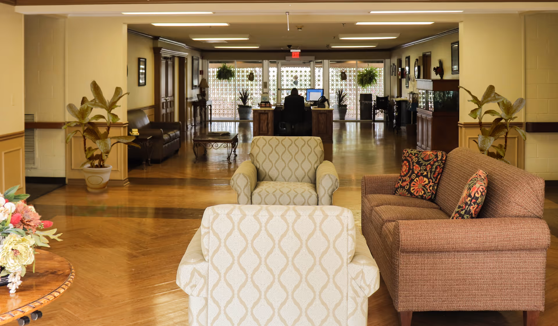 A spacious reception lounge with upholstered armchairs and a sofa facing a front desk and corridor with plants and wood floors.