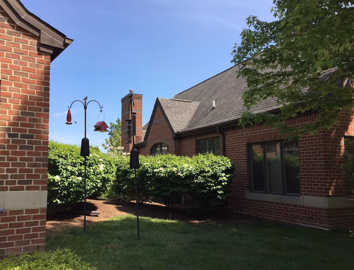 Outdoor view of a brick building with a sloped roof, surrounded by green bushes and trees under a clear blue sky. Two bird feeders are hanging on metal poles in the garden area.