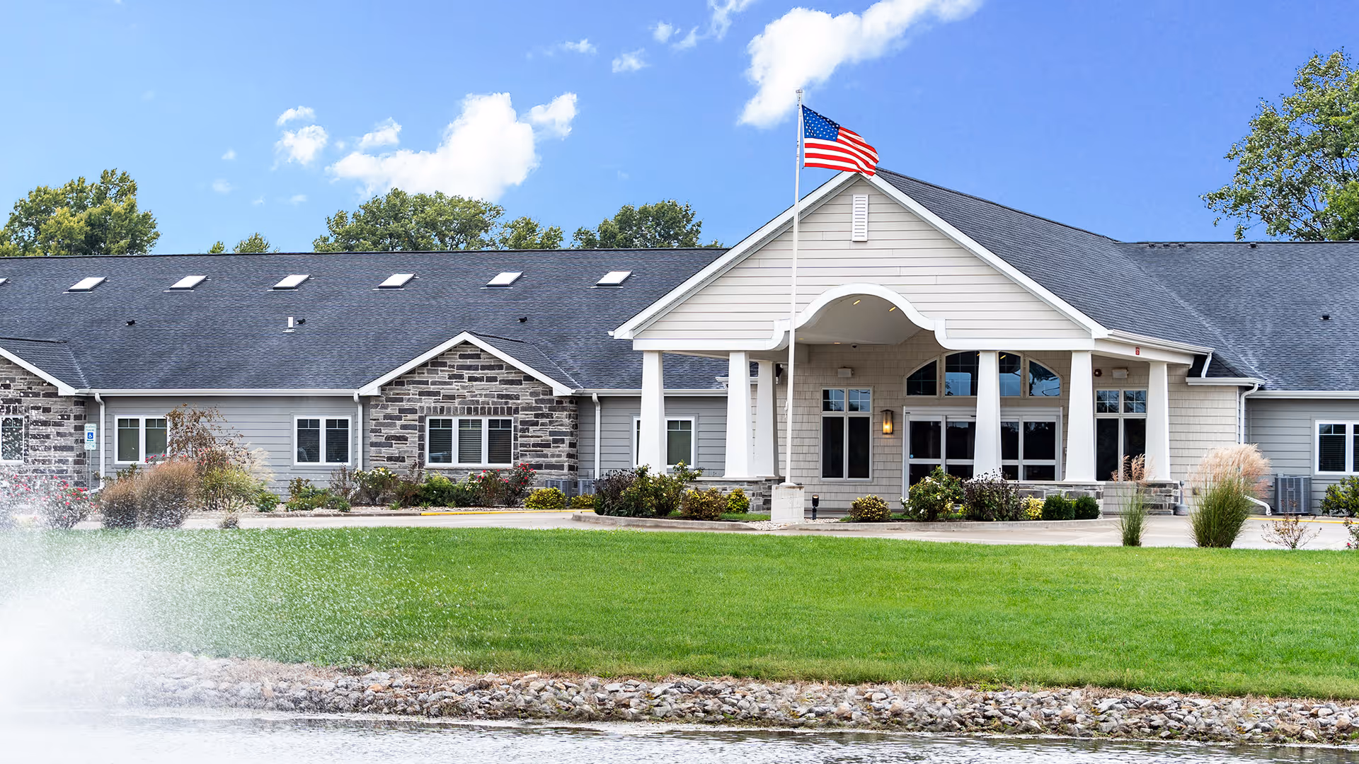 Front exterior view of a single-story senior living facility building with a peaked roof, stone and siding facade, an American flag on a flagpole, well-maintained green lawn, and a water fountain spraying in the foreground under a partly cloudy blue sky.
