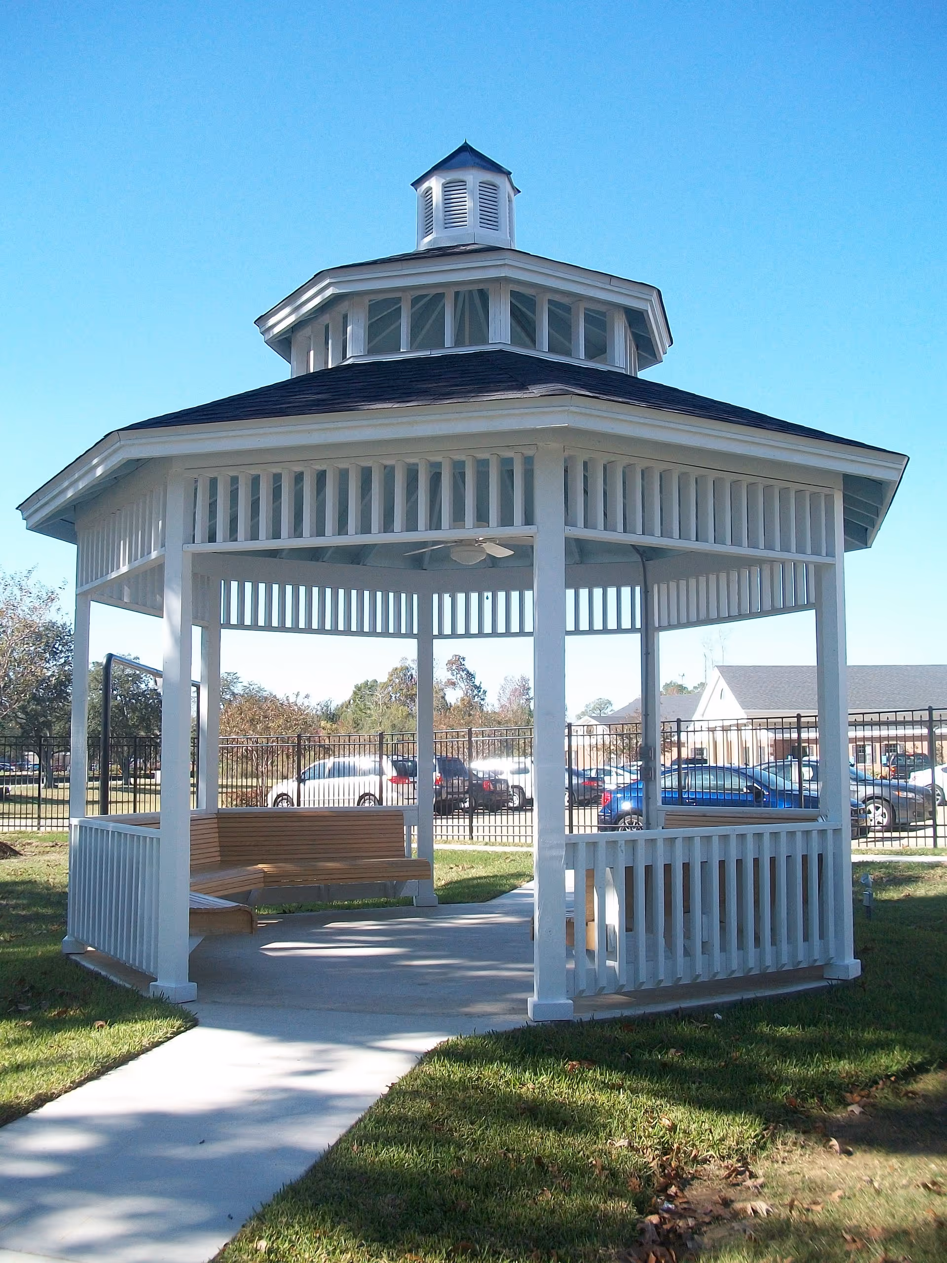 White wooden gazebo with a shingled roof and a small cupola on top, located outdoors on a grassy area with a concrete pathway leading to it. There are wooden benches inside the gazebo and a ceiling fan mounted on the roof. In the background, there is a black metal fence, parked cars, and some buildings under a clear blue sky.