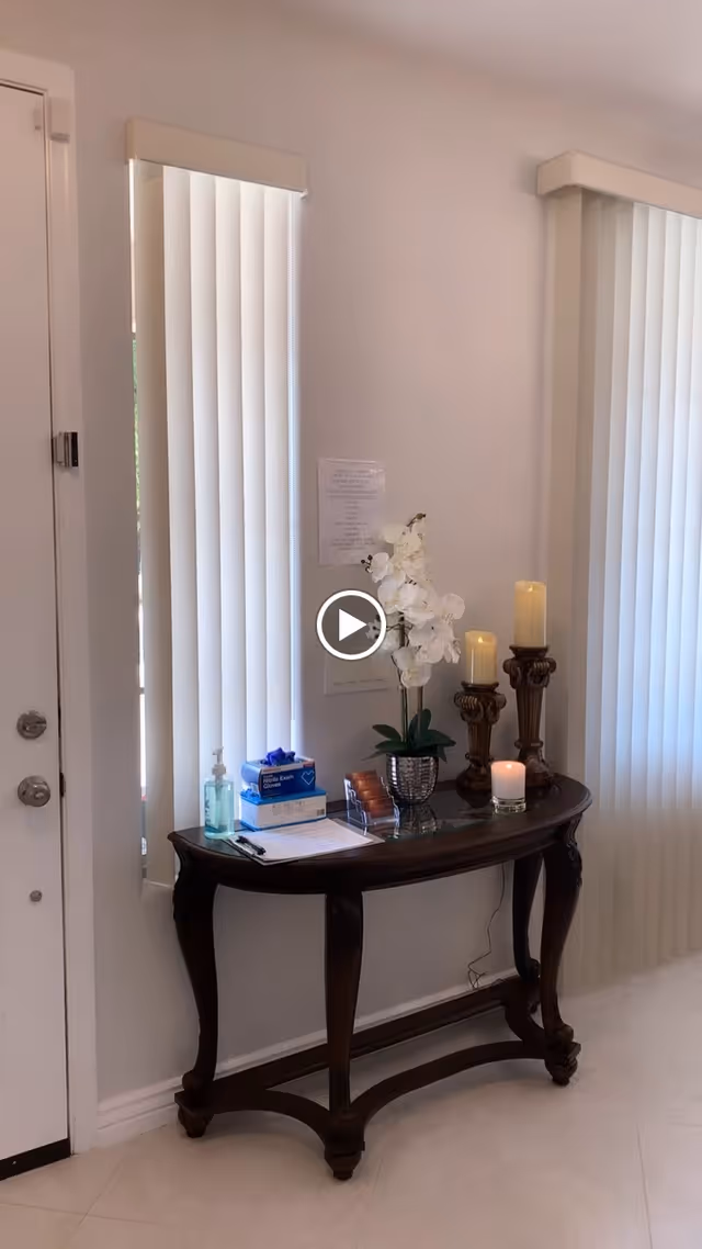 A wooden entry console table beneath two vertical-blind windows holding an orchid, candles, hand sanitizer, and paperwork.