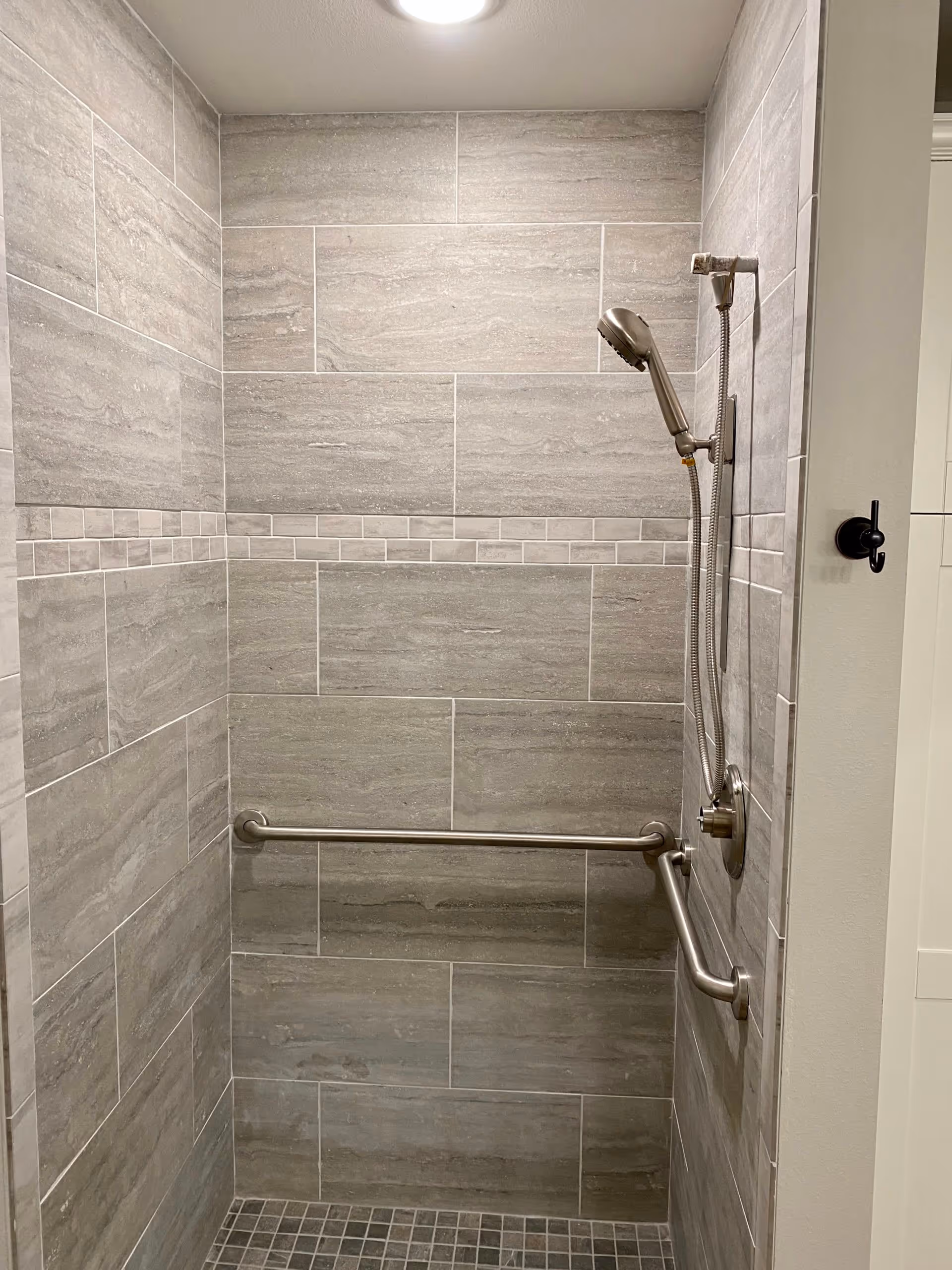 A tiled shower area with gray rectangular wall tiles and smaller square floor tiles. There is a horizontal grab bar on the back wall and an L-shaped grab bar on the right wall. A handheld showerhead is mounted on the right wall with a flexible hose. The shower area is well-lit with a ceiling light.