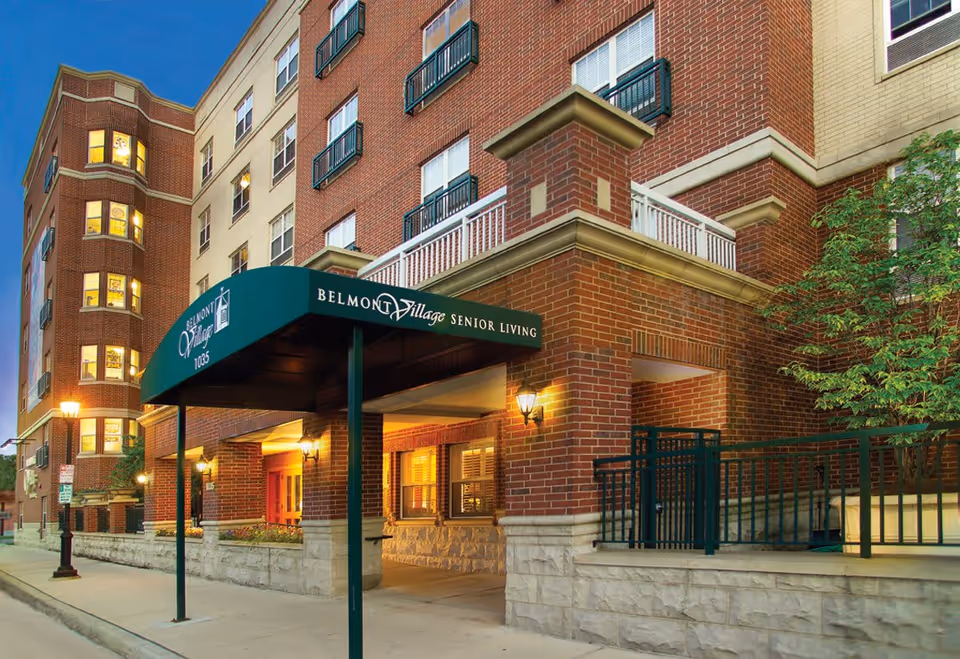 Exterior view of Belmont Village Senior Living Oak Park building at dusk, showing a multi-story brick facade with lit windows, a green awning with the facility name, and a sidewalk with street lamps.