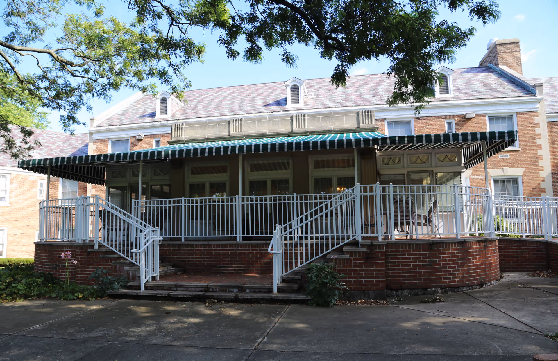 Exterior view of a brick building with a large covered porch featuring a green and white striped awning. The porch has white railings and stairs leading down to a concrete walkway. Trees partially shade the area in front of the building.