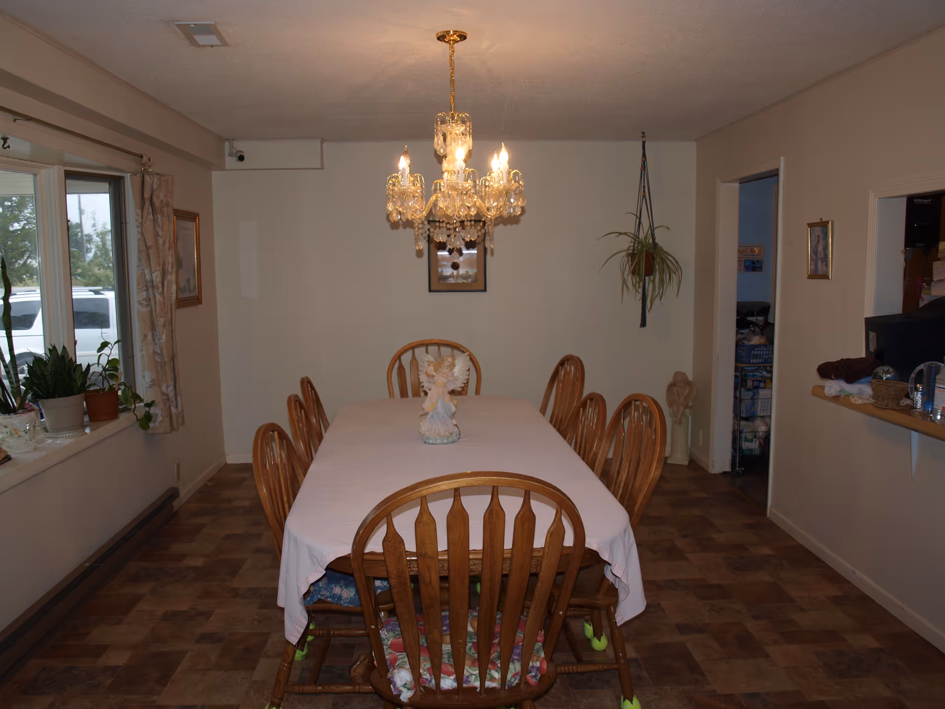 Dining room with a long rectangular wooden table covered with a white tablecloth and surrounded by eight wooden chairs with cushions. A crystal chandelier hangs above the table. There are plants on the windowsill to the left and a hanging plant on the right wall. The room has beige walls and a brown tiled floor.