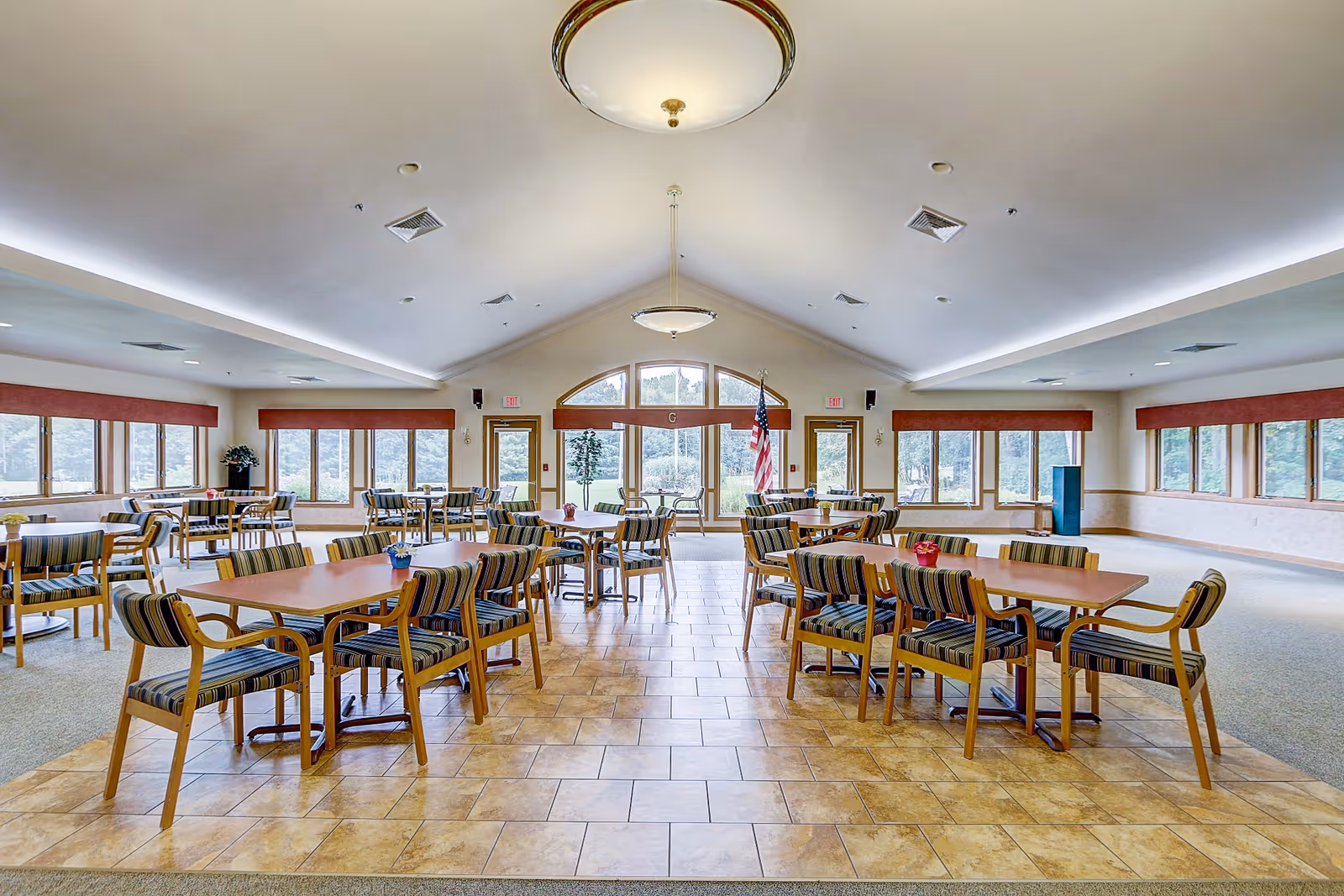 A spacious dining room with multiple wooden tables and striped cushioned chairs arranged neatly. Large windows with red valances line the walls, allowing natural light to fill the room. The floor is a combination of tiled and carpeted sections. There are two ceiling lights and an American flag near the back exit doors with glass panels.