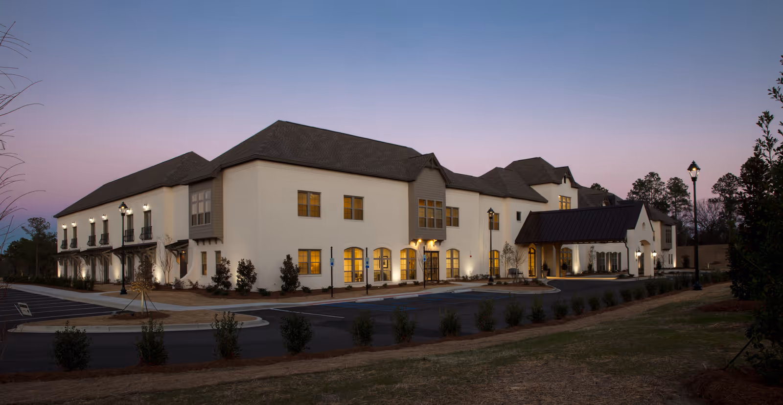 Front exterior of a large two-story residential building with lit windows, a covered entrance, and surrounding landscaping at dusk.
