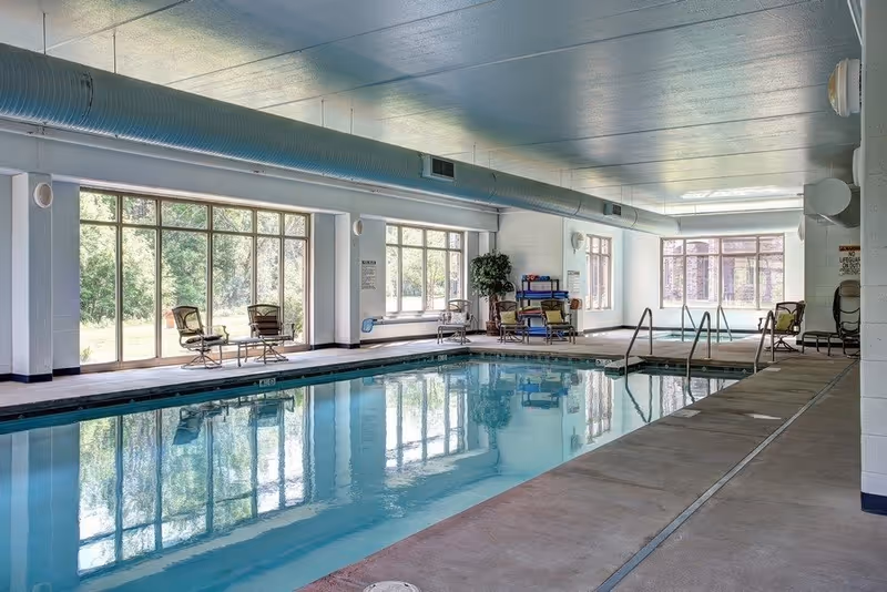 Indoor swimming pool area with large windows letting in natural light. Several chairs and small tables are arranged around the pool deck. The ceiling has exposed ductwork and the pool has metal handrails for entry and exit.