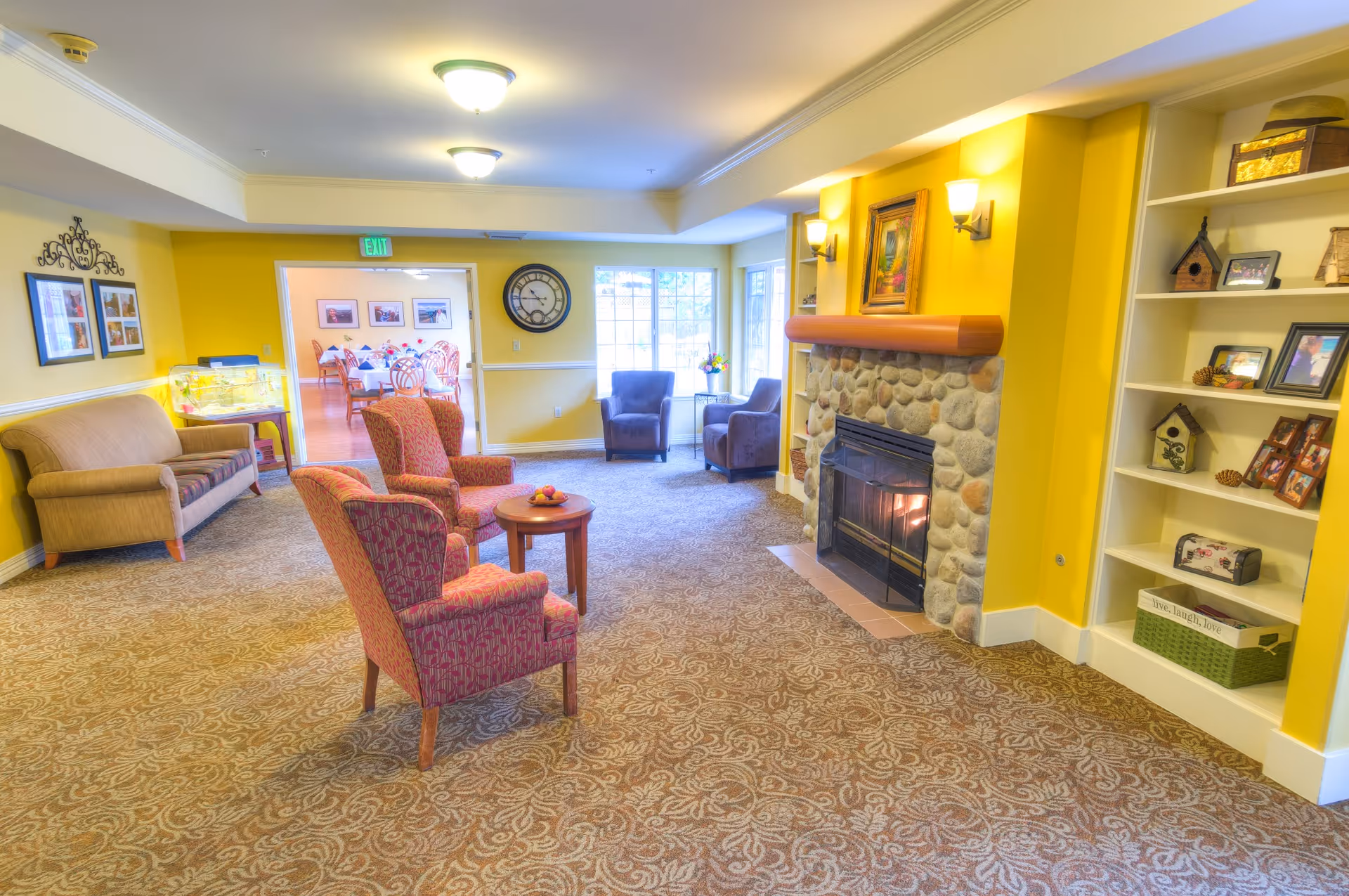 A cozy living room area with yellow walls, a stone fireplace with a fire burning, two red patterned armchairs around a small wooden table, a beige sofa, and two dark armchairs near large windows. There is a built-in white bookshelf with decorative items and framed photos. In the background, a dining room with tables and chairs is visible through an open doorway.