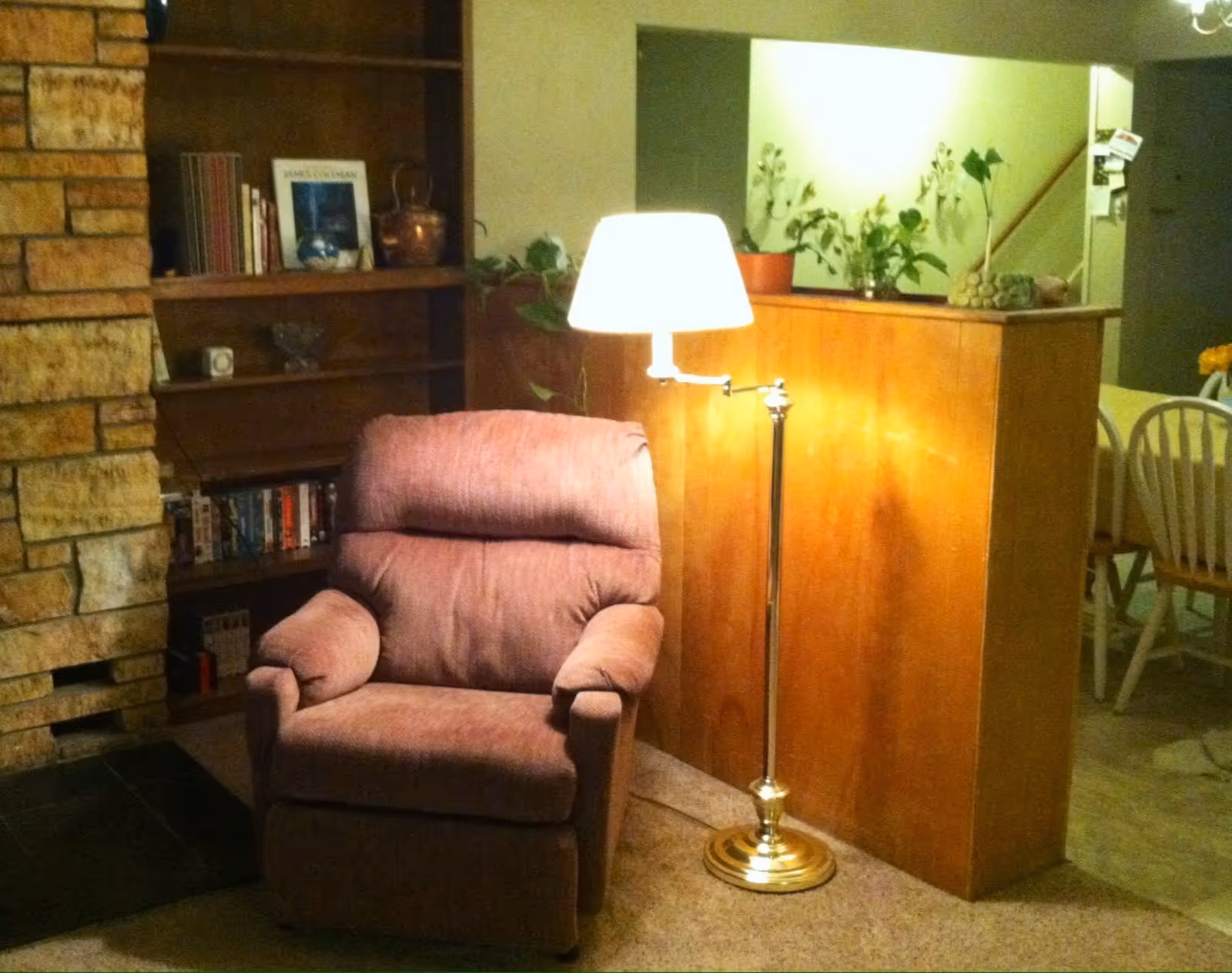 A cozy living room corner with a mauve recliner beside a brass floor lamp, bookshelf and a partial view of a dining area.