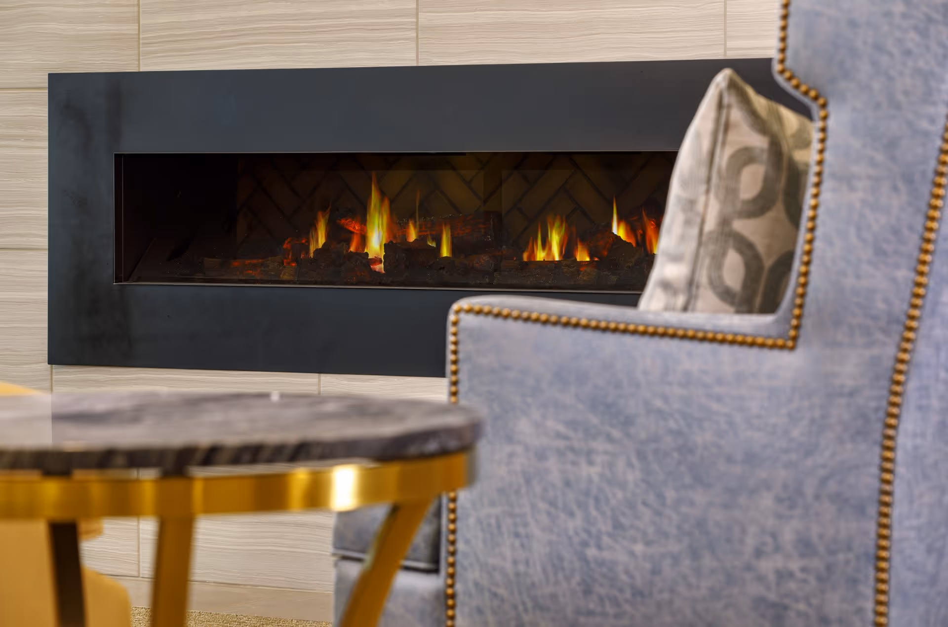 A modern indoor fireplace with visible flames and logs, set in a beige tiled wall. In the foreground, there is a blue upholstered chair with brass nailhead trim and a patterned cushion, along with a round table with a dark marble top and gold legs.