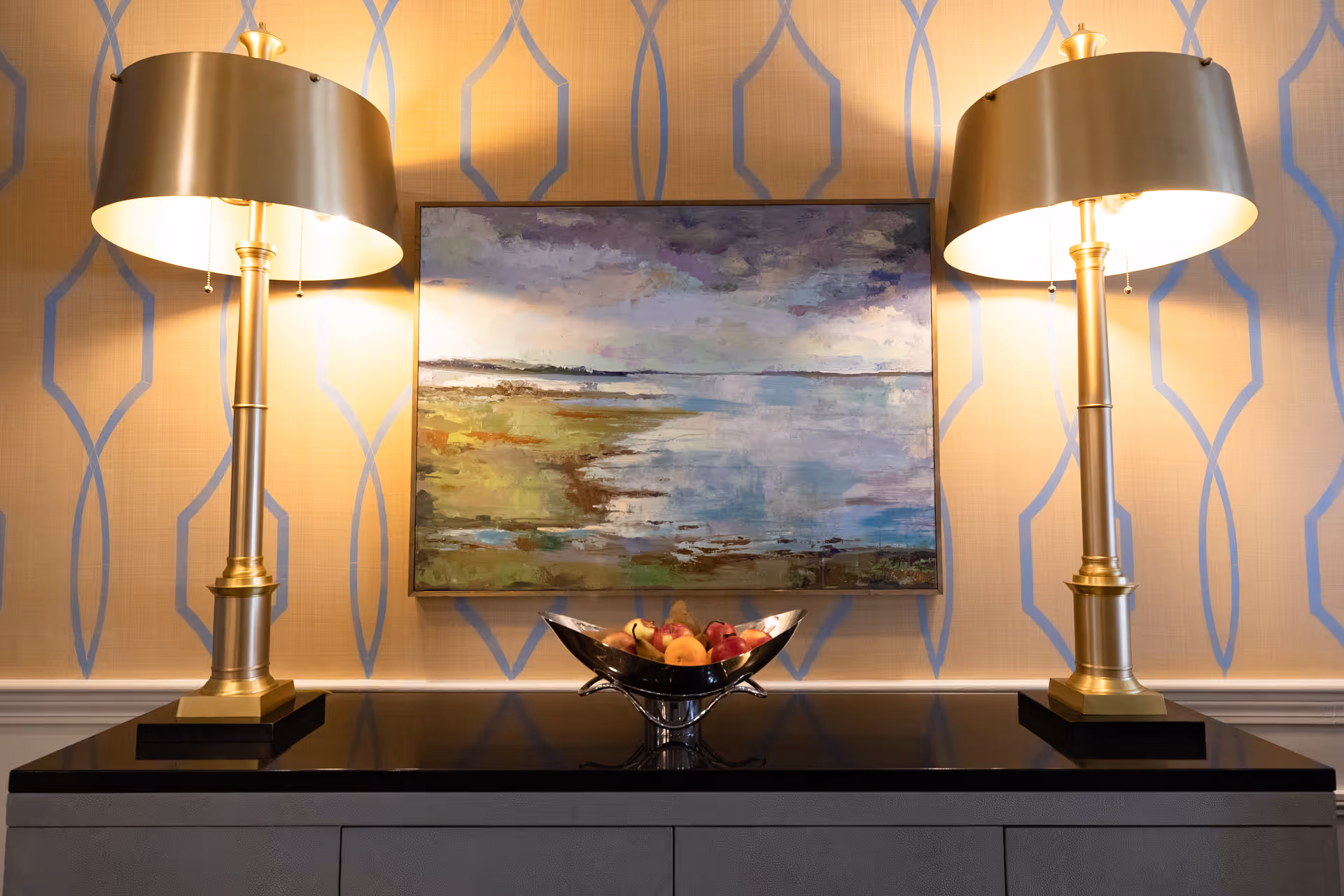 A decorative interior scene featuring a black countertop with a silver bowl filled with fruit in the center. On either side of the bowl are two gold-colored table lamps with black shades. Behind the countertop is a wall with patterned wallpaper and a framed landscape painting depicting a cloudy sky over a body of water.