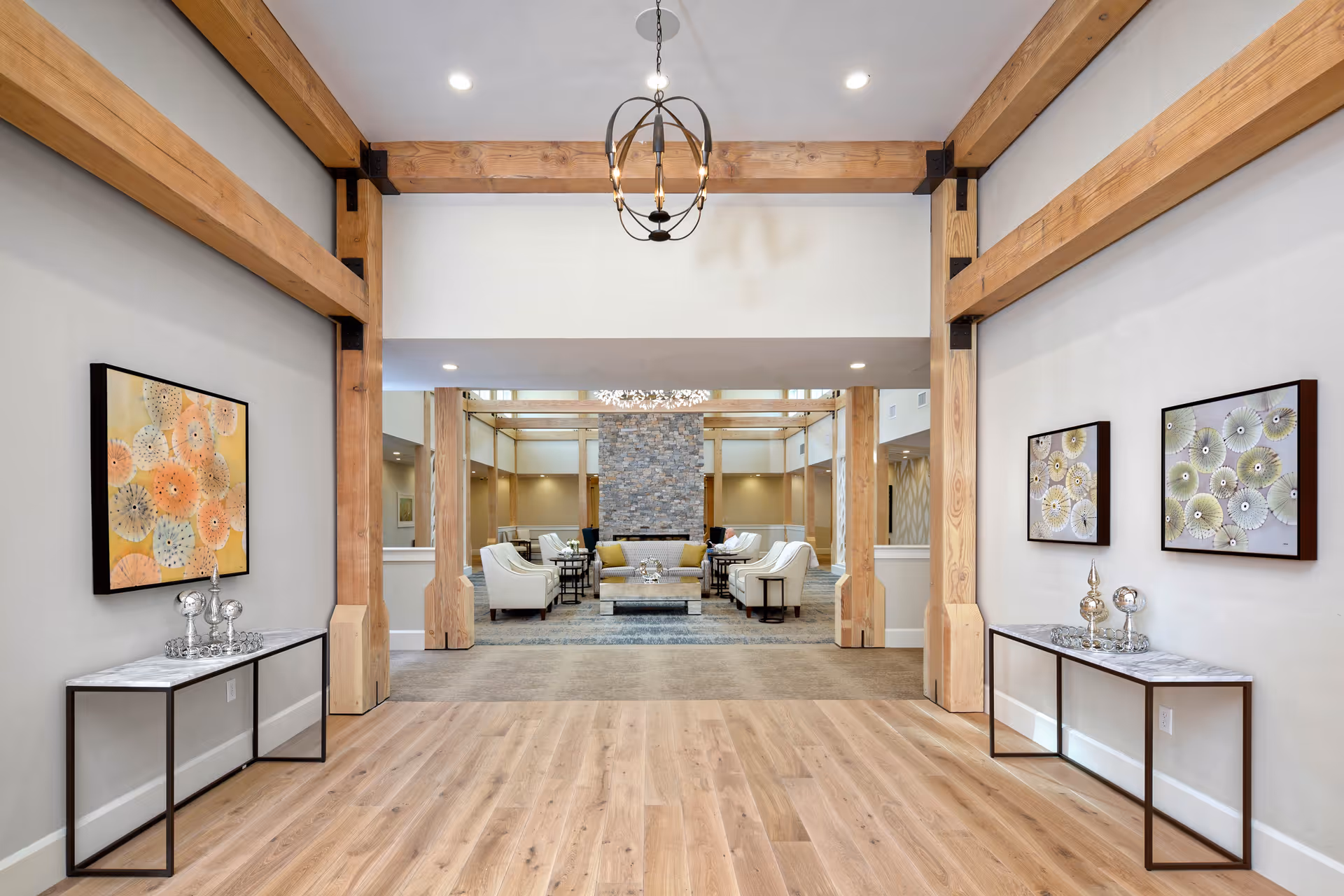 Wood beams frame a bright lobby with seating and artwork, leading to a stone fireplace lounge.