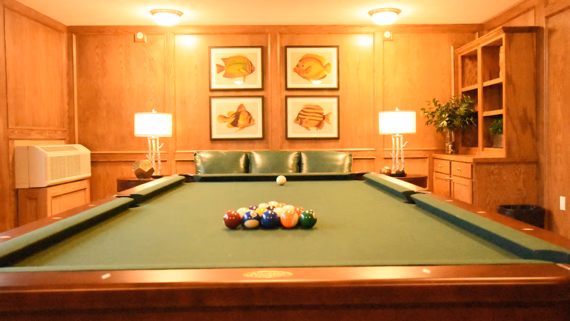 A pool table centered in a wood-paneled recreation room with framed fish art, lamps, and a green couch against the far wall.