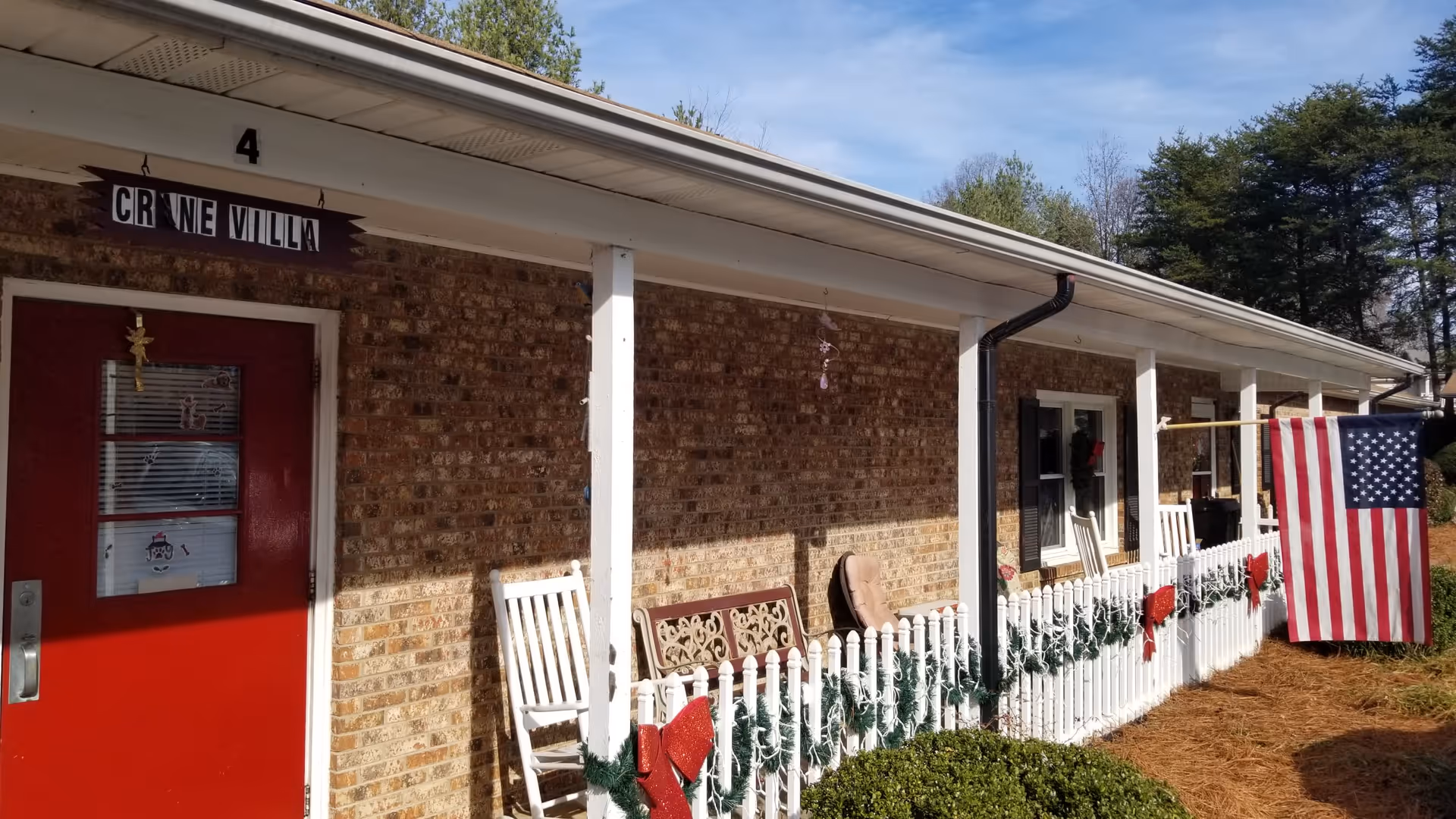 Exterior view of a brick building with a covered porch. The porch has white railings decorated with green garlands and red bows. There are white rocking chairs and a bench along the porch. An American flag is hanging on the right side. The door is red with a small window and a sign above it reads 'CRANE VILLA'.