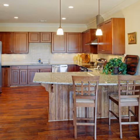 A spacious kitchen with wooden cabinets, a granite countertop island with two wooden chairs, hardwood flooring, and pendant lights hanging from the ceiling. The kitchen includes a stove, dishwasher, and decorative plants on the island.