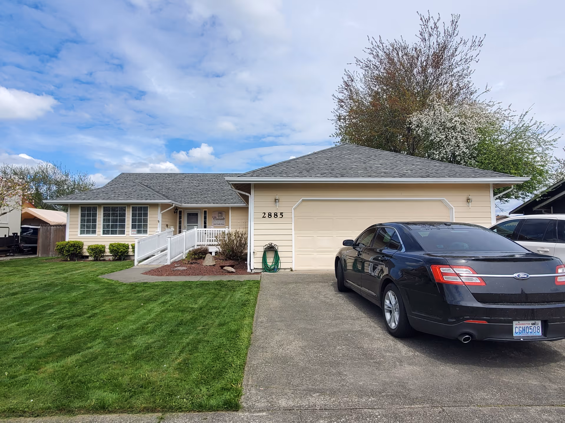 Front view of a single-story pale yellow house with an attached garage, driveway with a parked black car, green lawn, and a ramped entrance.