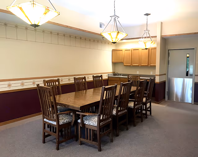 Communal dining room with a long wooden table surrounded by chairs, pendant lights overhead, and a small kitchenette and door in the background.