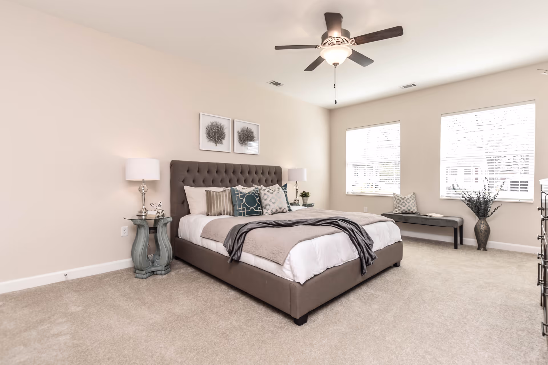 Spacious neutral bedroom with a tufted bed, nightstands, ceiling fan, two large windows and a bench.