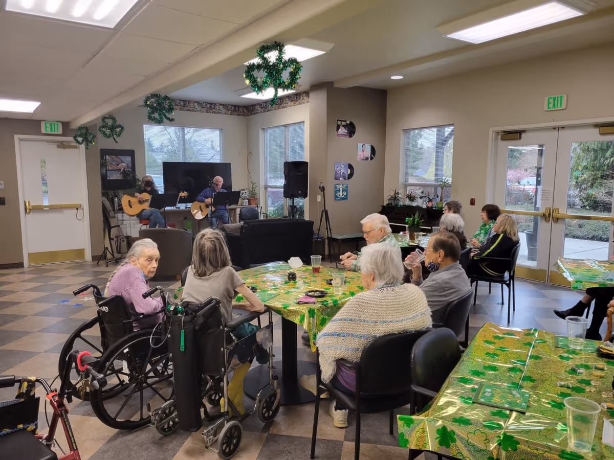 A group of elderly people seated around tables covered with green shamrock-themed tablecloths in a common room. Two musicians are playing guitars near a TV and speaker setup. The room has large windows and exit doors, with shamrock decorations hanging from the ceiling.