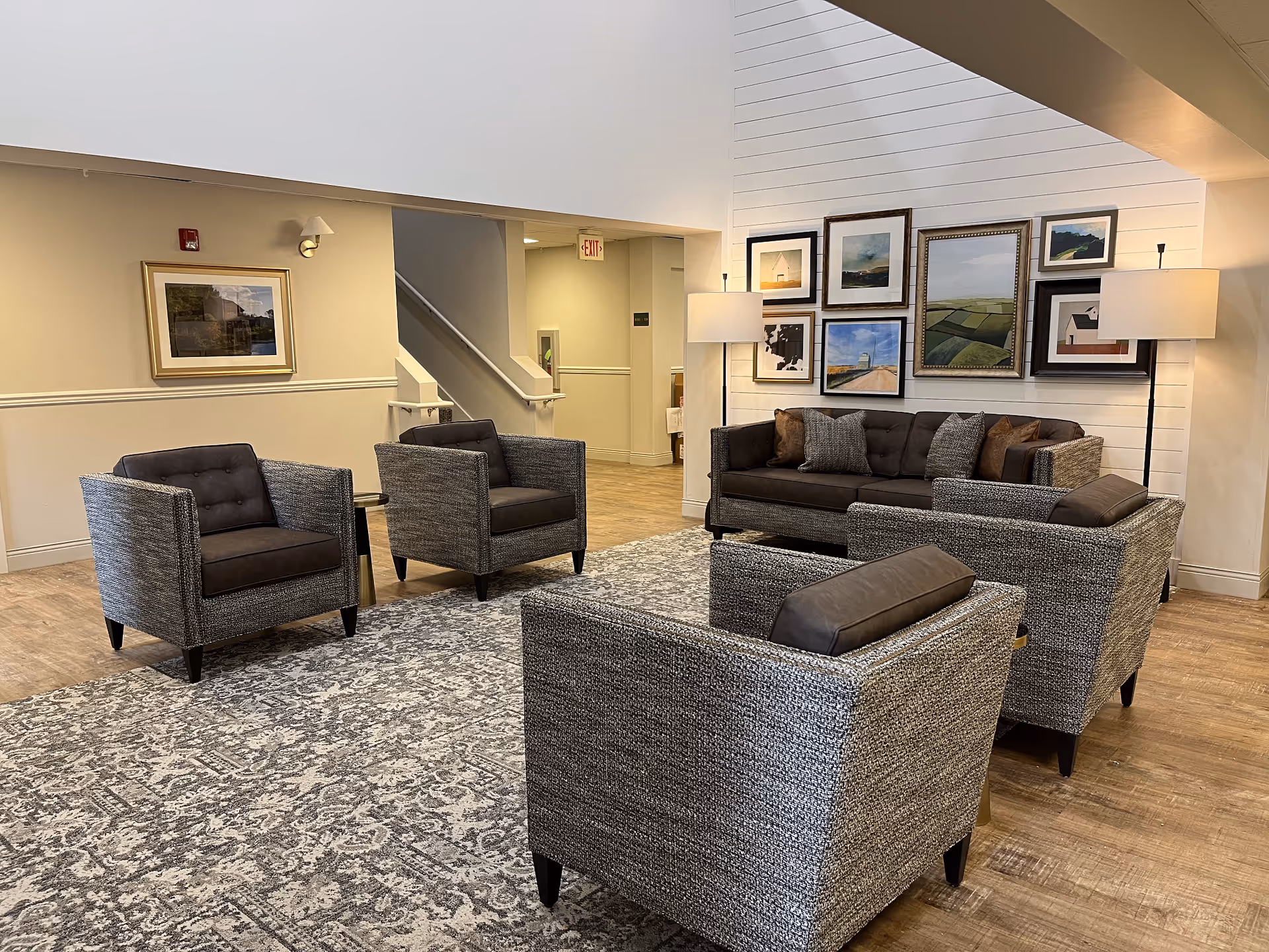 A cozy senior living facility lounge area with a gray patterned rug, four gray armchairs with dark cushions, and a dark gray sofa with multiple pillows. The walls are decorated with framed landscape paintings, and two floor lamps provide warm lighting. The floor is wooden, and there is a staircase and hallway visible in the background.