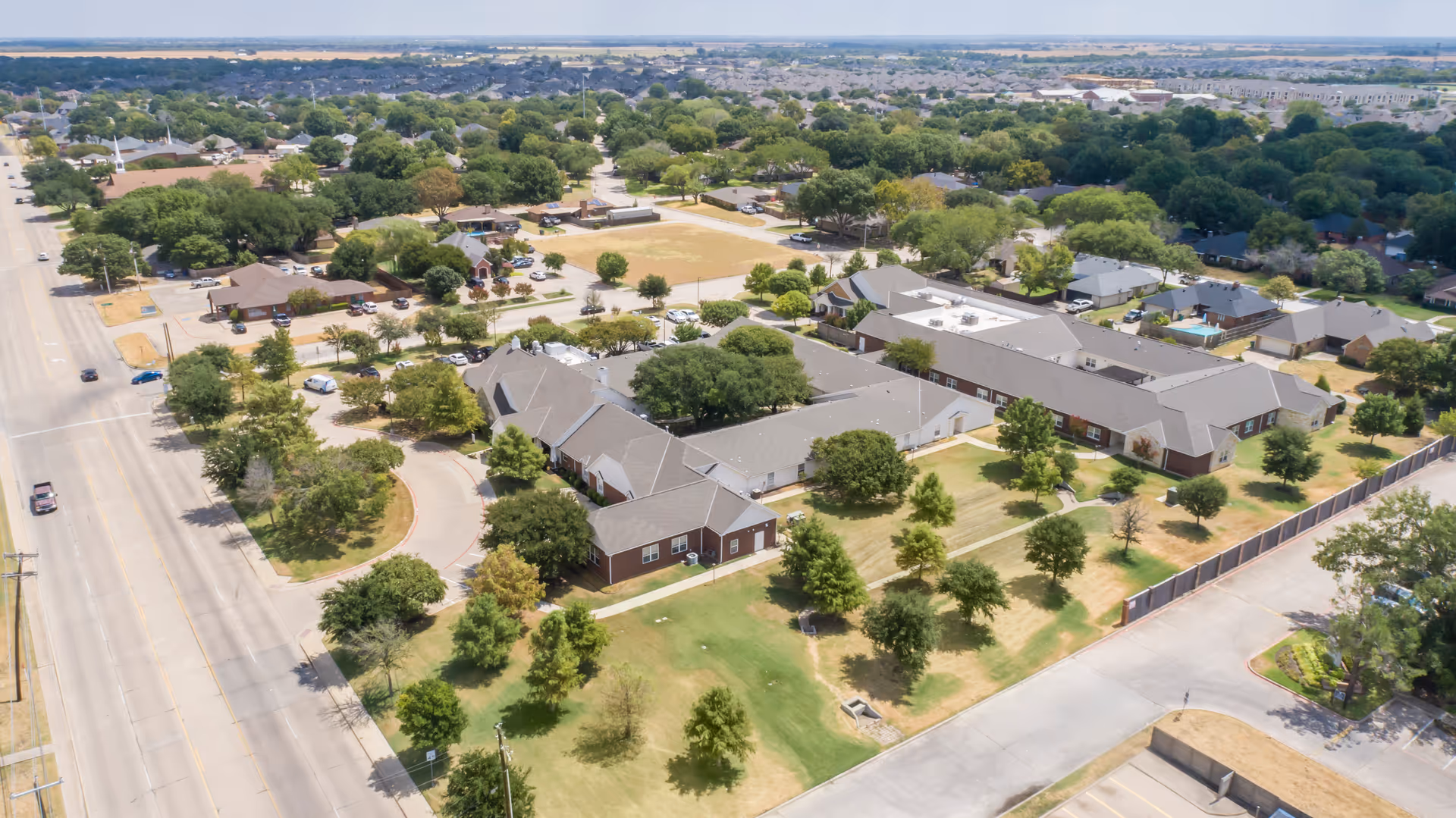Aerial view of Brookdale Waxahachie senior living facility showing multiple connected buildings with gray roofs surrounded by green lawns and trees. The facility is located in a suburban area with nearby residential houses and streets visible.