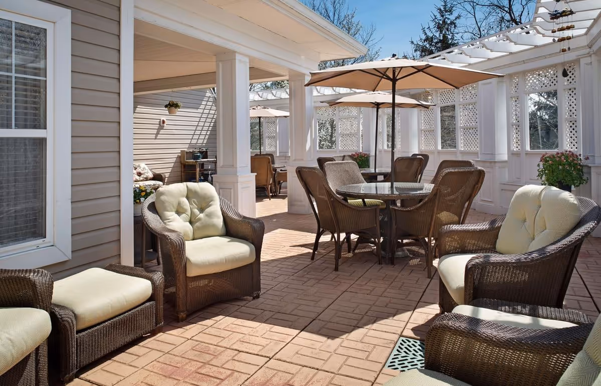 Outdoor patio area with cushioned wicker chairs and glass-top tables under large beige umbrellas, surrounded by white lattice walls and columns, with a clear blue sky in the background.